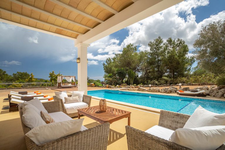 Piscina en villa de Ibiza con tumbonas y porche con sofás de exterior en día soleado